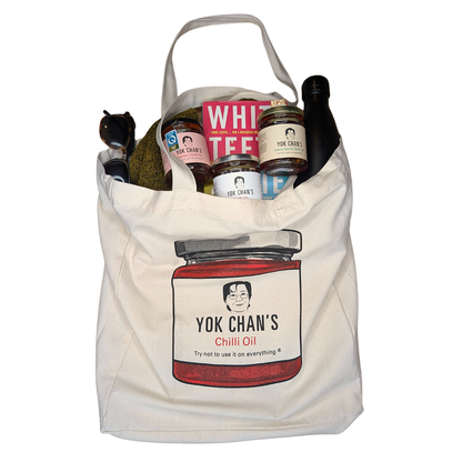 A canvas tote bag featuring a printed illustration of a jar labeled 'Yok Chan's Chilli Oil' with a cartoon face. The bag is filled with various items, including jars, a bottle, and a box, and is photographed against a plain white background.