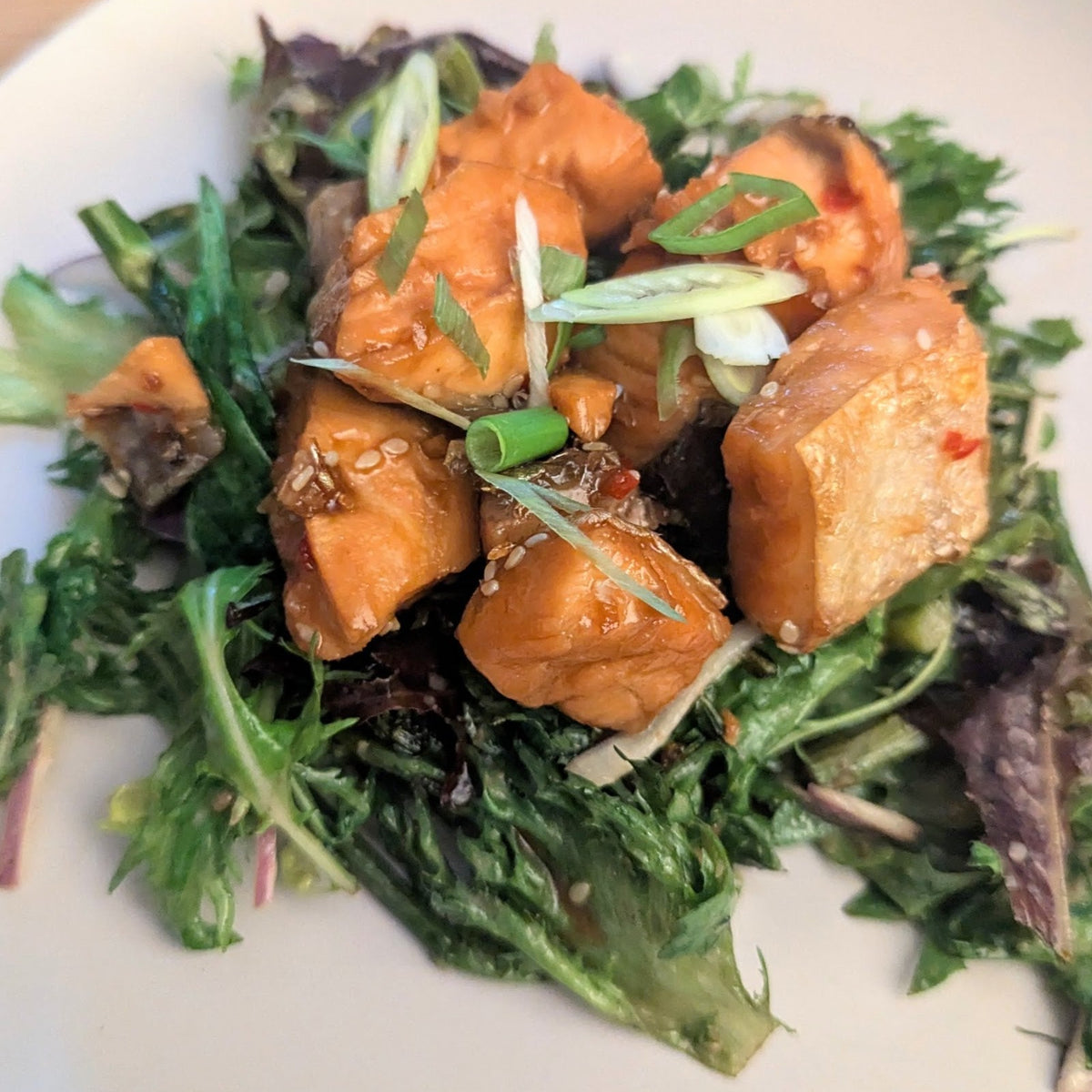 A close-up shot of a plated salad featuring glazed sweet potato cubes on a bed of mixed green and purple lettuce, garnished with sliced scallions, served on a white plate.