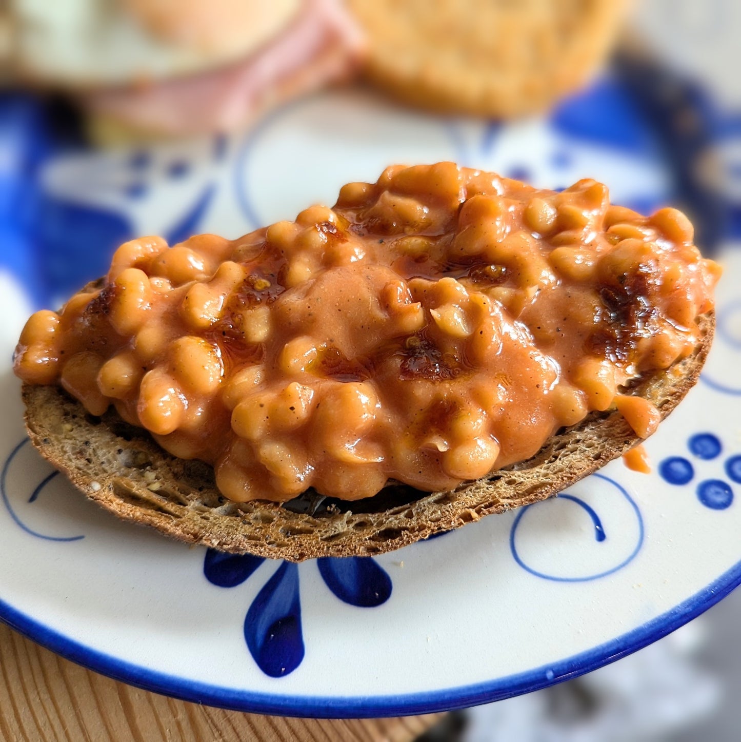 A close-up of baked beans spread over a slice of brown bread, resting on a blue and white patterned ceramic plate.