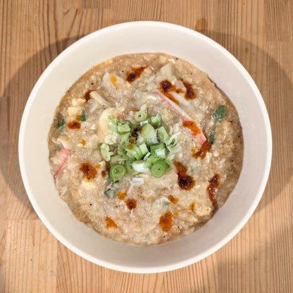 A top-down view of a bowl of oatmeal or porridge topped with chopped green onions, red onion, and hot sauce, served in a white bowl on a wooden table.
