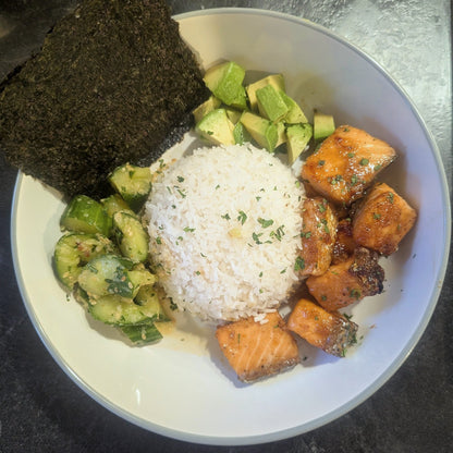 A top-down view of a white bowl containing a mound of white rice, cubed avocado, grilled salmon pieces, and a dark seaweed sheet, set on a dark countertop.