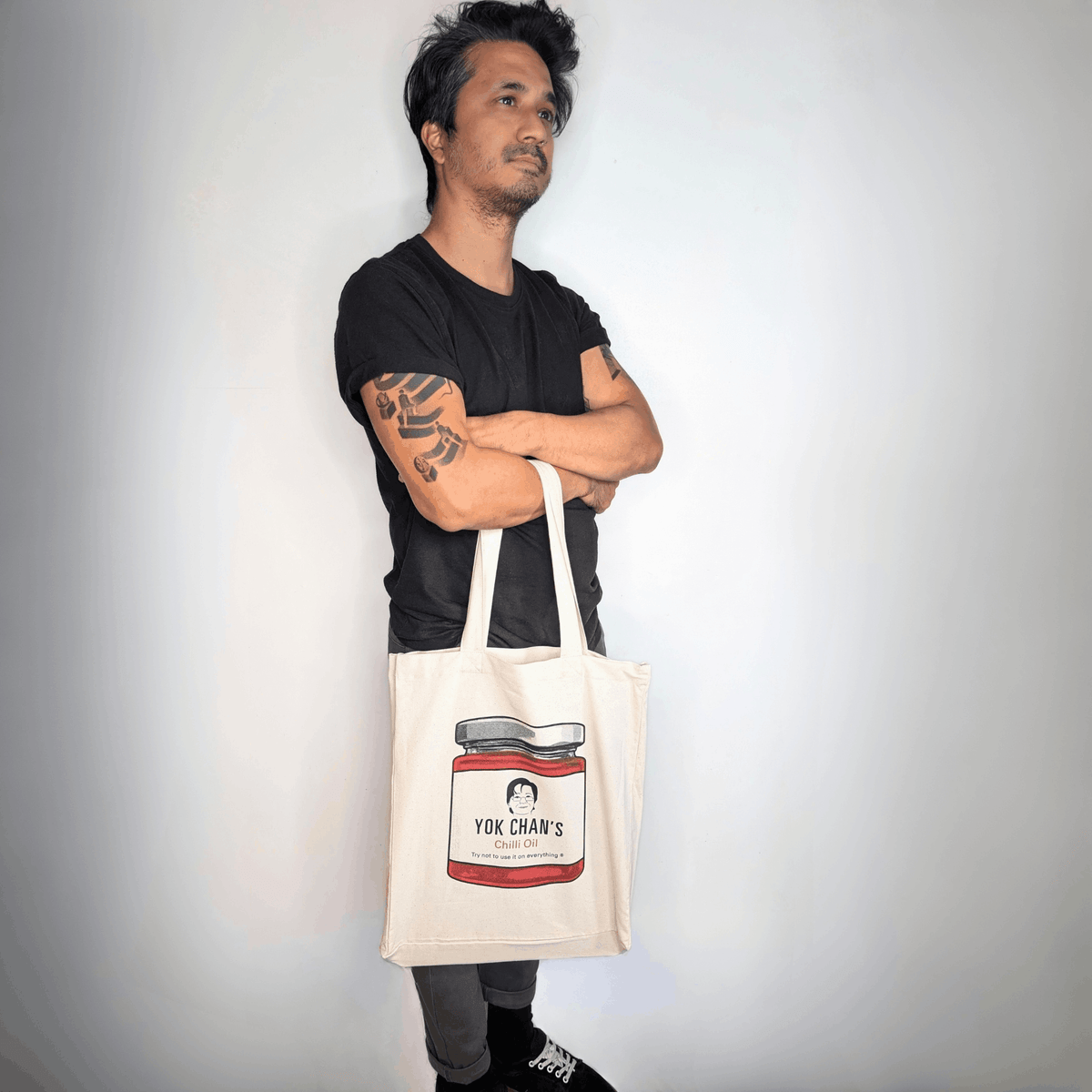 A man wearing a black t-shirt and grey pants stands with his arms crossed, holding a cream-coloured canvas tote bag. The tote bag features a printed illustration of a jar of Yok Chan's Chilli Oil