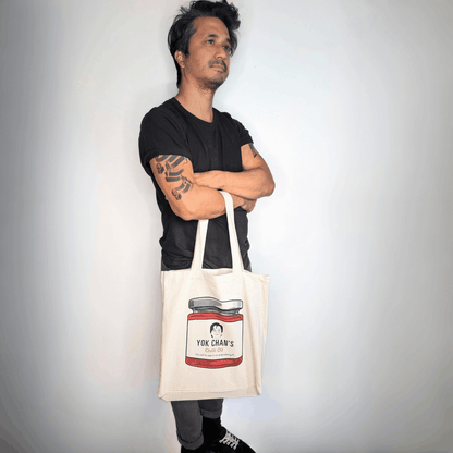 A man wearing a black t-shirt and grey pants stands with his arms crossed, holding a cream-coloured canvas tote bag. The tote bag features a printed illustration of a jar of Yok Chan's Chilli Oil