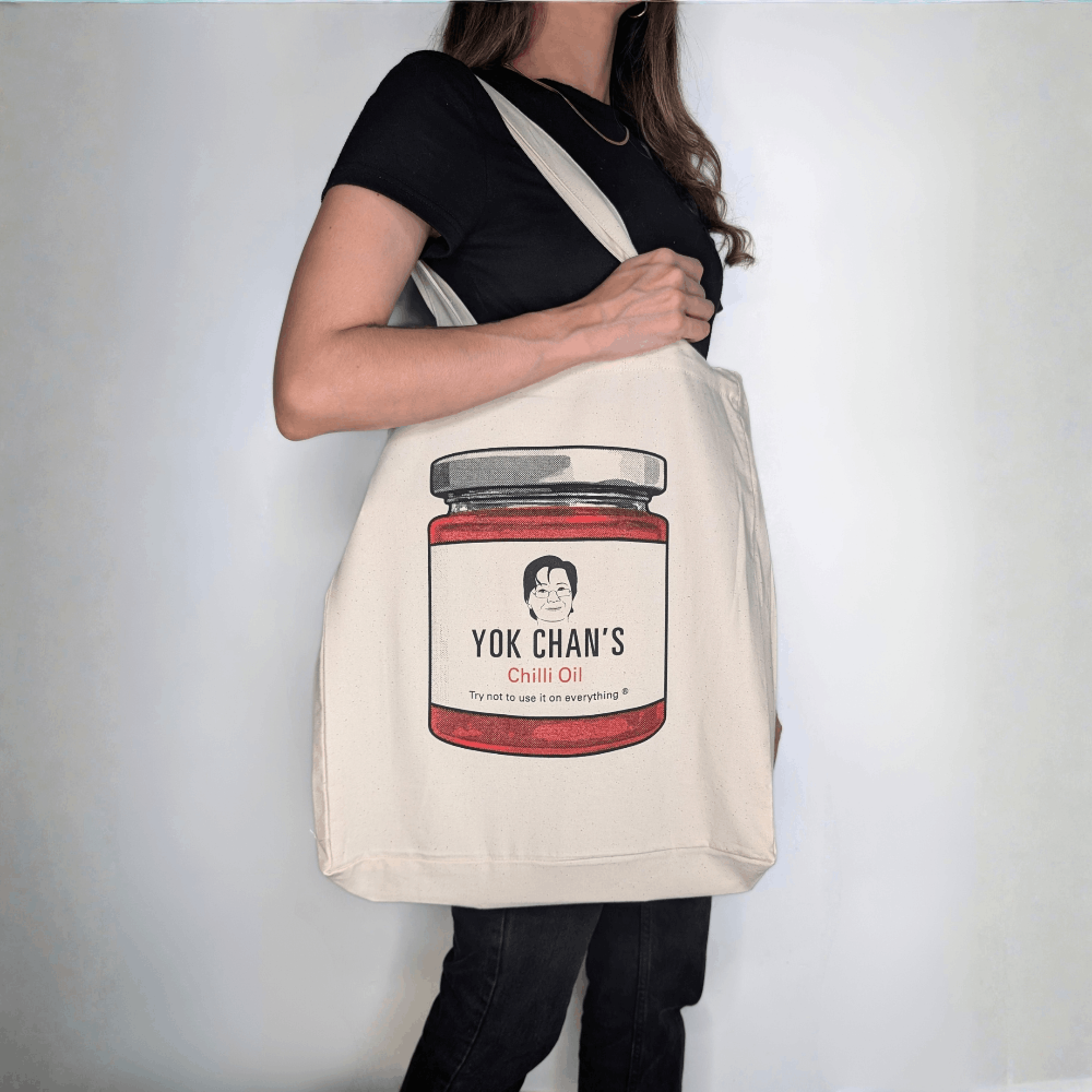 A woman wearing a black t-shirt and black jeans carries a cream canvas tote bag over her shoulder. The bag features a printed graphic of a red chilli oil jar labeled 'Yok Chan's Chilli Oil'.