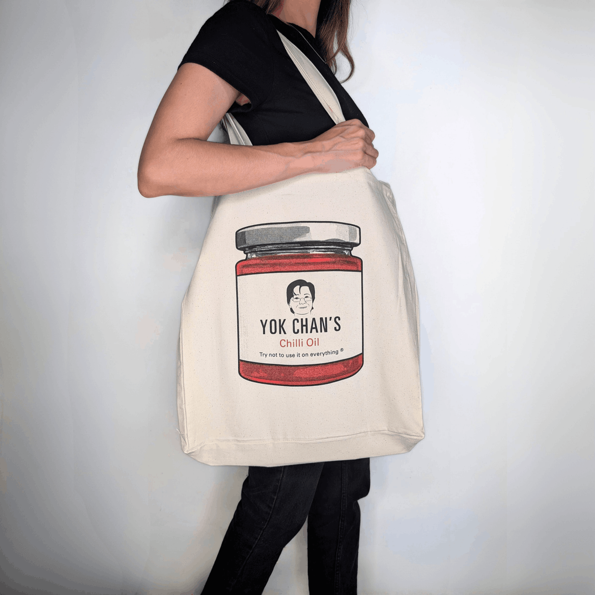 A person wearing a black shirt and black pants carries a large cream canvas tote bag with a printed illustration of a Yok Chan's Chilli Oil jar. The background is a plain white wall.