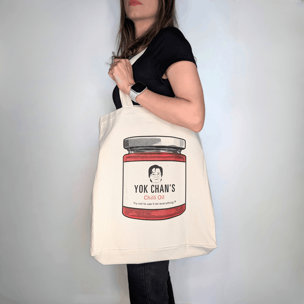 A woman wearing a black t-shirt and dark pants carries a cream canvas tote bag featuring a graphic of a Yok Chan's Chili Oil jar.