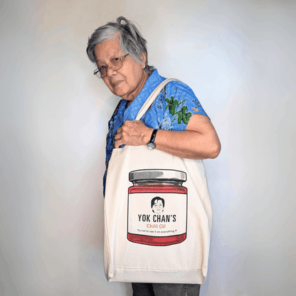 An older woman wearing glasses and a blue floral shirt carries a cream canvas tote bag featuring a printed graphic of a jar of Yok Chan's Chilli Oil.