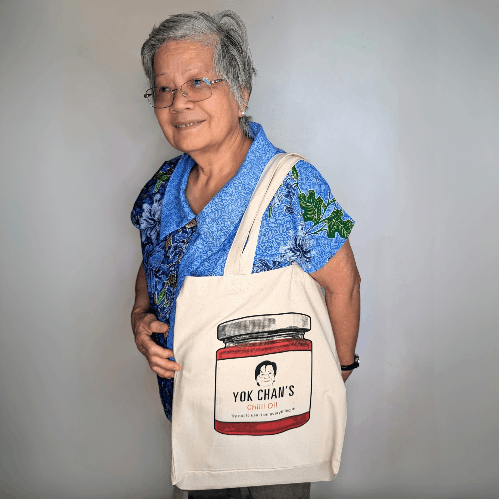 An elderly woman with gray hair and glasses is smiling while holding a cream canvas tote bag. The tote bag features a printed graphic of a red chili jar with the text 'YOK CHAN'S CHILI OIL'.