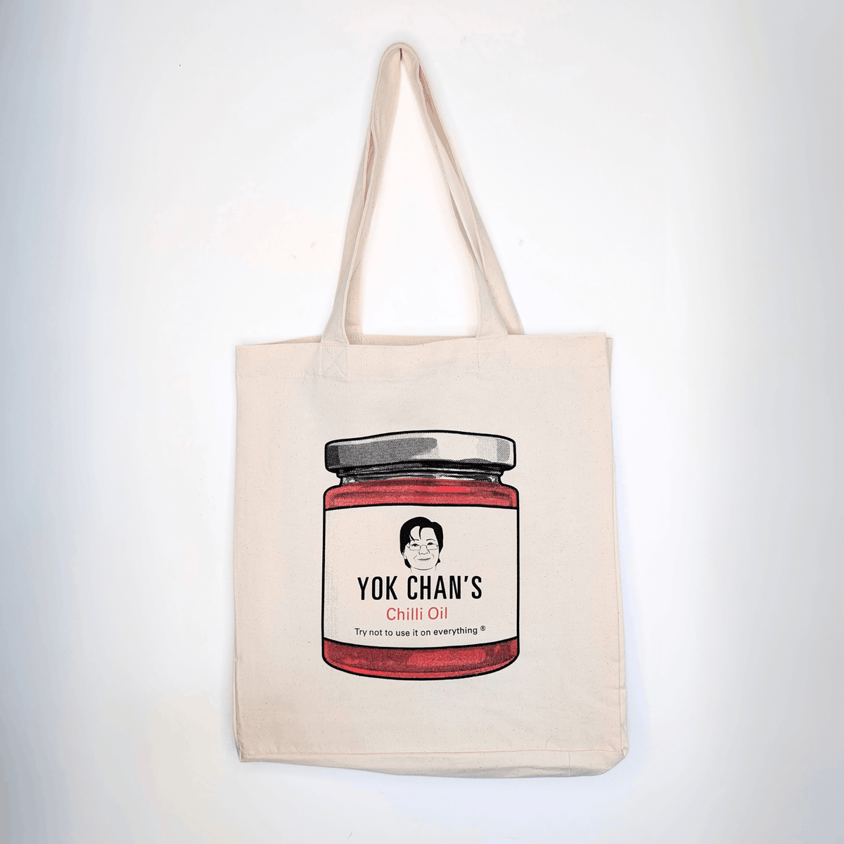 A beige canvas tote bag with a printed illustration of a jar of Yok Chan's Chili Oil on the front, hanging against a plain white wall.