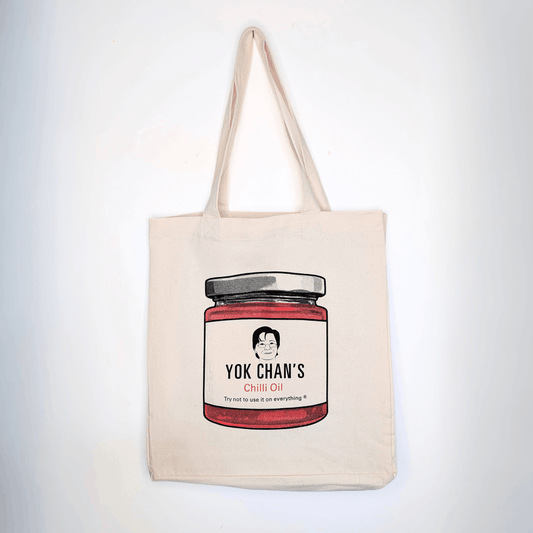 A beige canvas tote bag with a printed illustration of a jar of Yok Chan's Chili Oil on the front, hanging against a plain white wall.